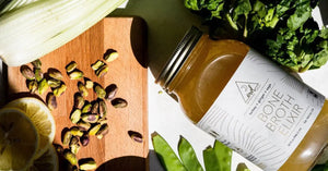 A jar of bone broth elixir with fresh vegetables, pistachios, and lemon slices on a wooden cutting board