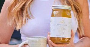 A woman holding a jar of vegan mineral broth elixir alongside a ceramic mug outdoors