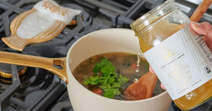 Bone broth being poured into a pot with herbs and vegetables for cooking