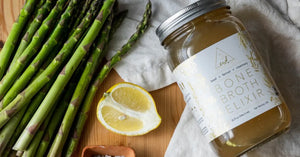 Bone Broth Elixir jar displayed with fresh asparagus, lemon, and sea salt on a wooden surface