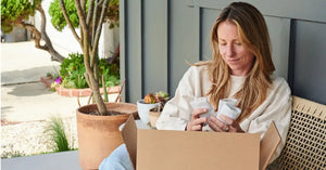 Woman unpacking bone broth items from a box while sitting on a porch surrounded by potted plants