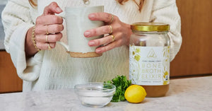 Bone Broth Elixir jar beside fresh parsley, lemon, and salt, with a ceramic mug being prepared for serving
