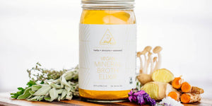 Vegan bone broth elixir in a glass jar surrounded by fresh herbs, mushrooms, ginger, & turmeric on a wooden surface