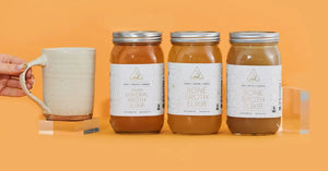 Premium bone broth and vegan elixirs arranged against an orange background with a hand holding a ceramic mug.