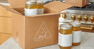 Organic bone broth jars neatly packed in a branded cardboard box, displayed on a kitchen counter with a stove in the background.