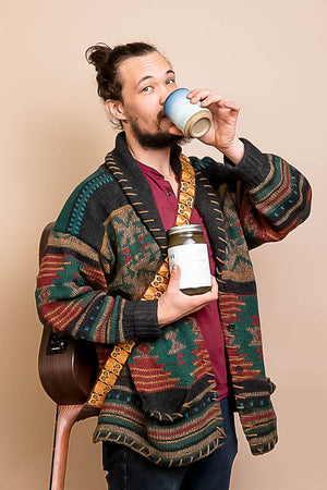 Man sipping from a mug & holding a jar of bone broth, wearing a colorful patterned sweater against a beige background