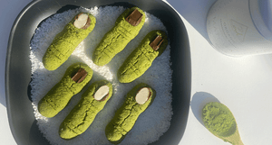 Matcha dessert shaped like green fingers with almond and chocolate details, served on shredded coconut in a black tray