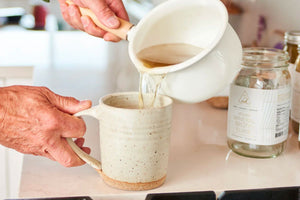 Bone broth cleanse being prepared as warm broth is poured from a white pot into a ceramic mug on a kitchen counter
