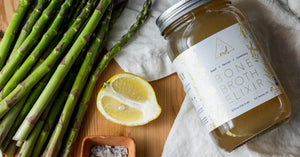 Natural bone broth jar labeled "Bone Broth Elixir" paired with fresh asparagus, lemon, and coarse salt on a wooden surface.