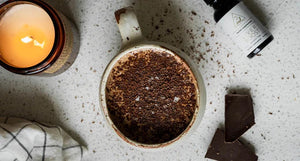 Dandelion Cocoa Latte in a rustic mug topped with grated chocolate and sea salt, surrounded by dark chocolate, a lit candle, and OWL Liver Boost herbal supplement
