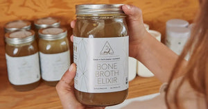 Delicious bone broth elixir held in hands with jars of broth stacked on shelf in background.