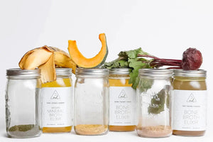 Bone broth nutrition jars and ingredients including beets, squash, and herbs displayed in a clean, white setting.