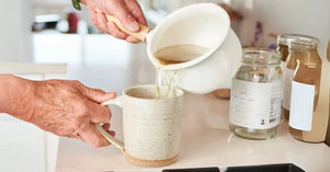 Organic bone broth being poured from a white saucepan into a ceramic mug beside an empty vegan mineral broth jar