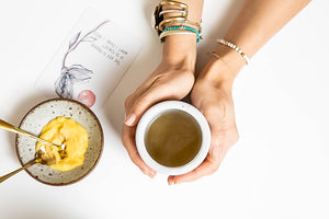 Hands holding a mug of bone broth next to a bowl of golden paste with spoons, placed on a white surface